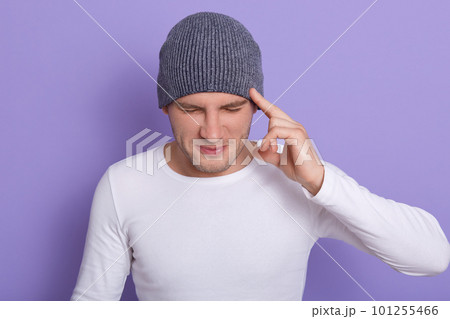 Studio shot of attractive male with closed eyes, keeps finger on temples, has terrible headache or forget something important, wearing white casual shirt and gray cap, isolated over lilac background. 101255466