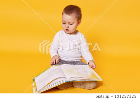 Toddler girl pretends to read book while sitting on floor, viewing pictures and turning pages, little girl looks concentrated, wearing casual white shirt, posing isolated over yellow background. Toddler girl pretends to read book while sitting on floor, viewing pictures and turning pages, little girl looks concentrated, wearing casual white shirt, posing isolated over yellow background. 101257325