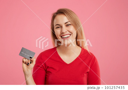 Isolated shot of cheerful pretty female holds plastic card in hands, wears red sweater, going to make online purchases, glad to recieve money bonus on her account, poses against pink background 101257435