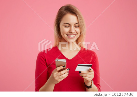 Happy beautiful woman holds smart phone and credit card, makes online payment, pays for purchase in online shop or verifies her bank account, isolated over pink background. Technology and accounting 101257436