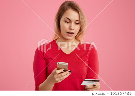 Shot of puzzled young woman with light short hair, wears red sweater, holds mobile phone and credit card, connected to wireless internet, makes shopping online, has surprised facial expression Shot of puzzled young woman with light short hair, wears red sweater, holds mobile phone and credit card, connected to wireless internet, makes shopping online, has surprised facial expression 101257439