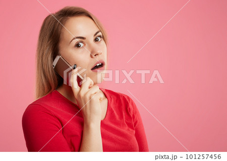 Shocked female recieves telephone call, speaks with stranger, hold smart phone and looks with surprised terrfied expression into camera, isolated over pink background with copy space for your text Shocked female recieves telephone call, speaks with stranger, hold smart phone and looks with surprised terrfied expression into camera, isolated over pink background with copy space for your text 101257456