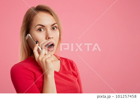 Shocked puzzled young female has phone conversation via telephone, surprised to hear unexpected news, isolated over pink background with copy space for your advertisment or promotional text. Shocked puzzled young female has phone conversation via telephone, surprised to hear unexpected news, isolated over pink background with copy space for your advertisment or promotional text. 101257458