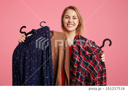 Happy lovely young woman holds clothes on hangers, rejoices new purchase, doesn`t know what to choose, going to spend money on new outfit, isolated over pink studio background. Shopping concept 101257516