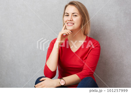 Indoor shot of pleasant looking happy female with pleasant smile on face, dressed in red sweater, poses at camera against grey background. Beautiful woman being in good mood after date with lover Indoor shot of pleasant looking happy female with pleasant smile on face, dressed in red sweater, poses at camera against grey background. Beautiful woman being in good mood after date with lover 101257999