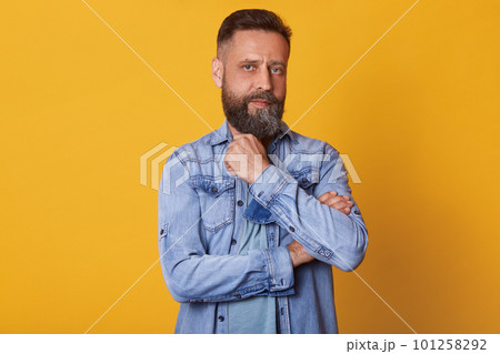 Strong thoughtful blue eyed man looking directly at camera, posing isolated over bright yellow background in studio, touching his beard with fingers, having powerful look, wearing in casual manner. Strong thoughtful blue eyed man looking directly at camera, posing isolated over bright yellow background in studio, touching his beard with fingers, having powerful look, wearing in casual manner. 101258292