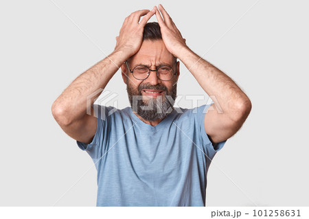 Bearded man in gray t shirt, has round eyewear, keeps hand on head, grimaced, has problems, being in bad mood, has terrible headache, being in conflict wit his wife. People emocions concept. Bearded man in gray t shirt, has round eyewear, keeps hand on head, grimaced, has problems, being in bad mood, has terrible headache, being in conflict wit his wife. People emocions concept. 101258631