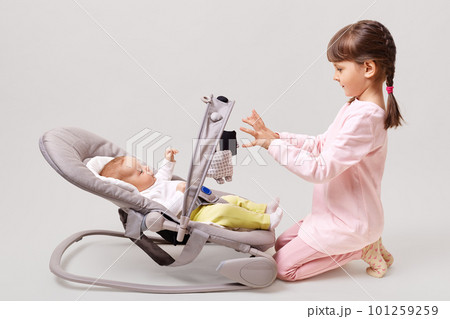 Profile of cute little girl with pigtails wearing pink casual clothing playing with new born baby sister girl lying in bouncer chair play, isolated over white background. Profile of cute little girl with pigtails wearing pink casual clothing playing with new born baby sister girl lying in bouncer chair play, isolated over white background. 101259259