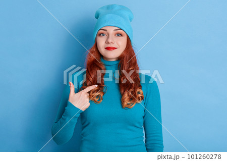 Satisfied Caucasian woman with red hair points at herself, stands self confident, dressed casual sweater and cap, feels successful of her own achievement, stands indoor against blue background. 101260278