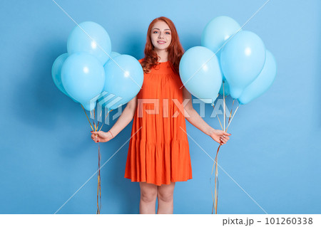 Young pretty woman with two bunches of balloons standing against blue wall and looking smiling directly at camera, celebrating holiday, girl with red hair wearing dress . Young pretty woman with two bunches of balloons standing against blue wall and looking smiling directly at camera, celebrating holiday, girl with red hair wearing dress . 101260338