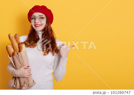 Happy Parisian girl with positive smile indicates aside at blank space, isolated over yellow background, French woman in red beret and spectacles has broad smile, stands with long bread baguettes. Happy Parisian girl with positive smile indicates aside at blank space, isolated over yellow background, French woman in red beret and spectacles has broad smile, stands with long bread baguettes. 101260747