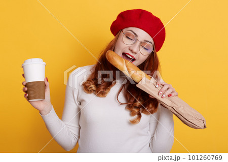 Portrait happy hungry woman holding paper bag with long white bread baguette and biting it, stands with coffee to go, wearing red beret and glasses, isolated over yellow background. Portrait happy hungry woman holding paper bag with long white bread baguette and biting it, stands with coffee to go, wearing red beret and glasses, isolated over yellow background. 101260769