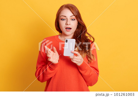 Stunned European woman being in shock, reads terrible news from cellular gadget, has wavy red hair, wears orange sweater, poses against yellow wall. 101261003
