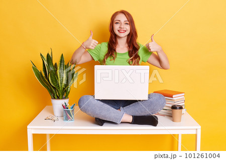 Happy excited lady wearing green t shirt and jeans, sitting on table with laptop on knees, sits ear stack of books and flower pot, female with red lips showing thumbs up, being in high spirit. Happy excited lady wearing green t shirt and jeans, sitting on table with laptop on knees, sits ear stack of books and flower pot, female with red lips showing thumbs up, being in high spirit. 101261004