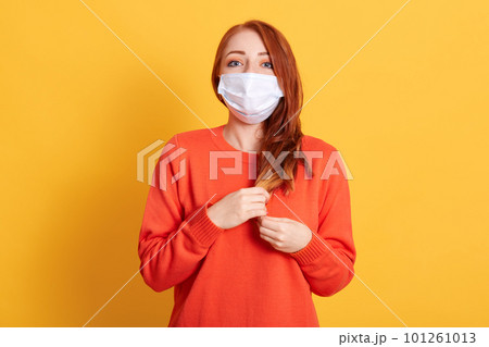 Gorgeous red haired young female wearing protective mask against COVID virus, tired of stress, looks confidently at camera with fear in eyes, poses against yellow background. 101261013