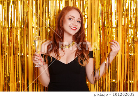 Portrait of girl with glass of champagne, posing against wall decorated with golden tinsel, lady wearing black dress and necklace., red haired smiling female celebrating holiday. Portrait of girl with glass of champagne, posing against wall decorated with golden tinsel, lady wearing black dress and necklace., red haired smiling female celebrating holiday. 101262035