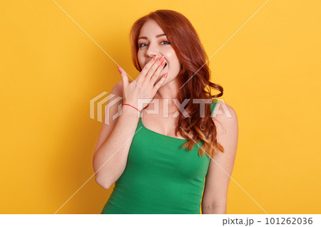 Beautiful red haired woman yawns isolated over yellow background, wears casual green t shirt, keeps palm near mouth, pleasant looking female looks at camera. Beautiful red haired woman yawns isolated over yellow background, wears casual green t shirt, keeps palm near mouth, pleasant looking female looks at camera. 101262036