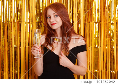 Lady with pleasant appearance looking directly at camera with glass of wine in hands, looking at camera, keeping hand in her hair, wearing black dress. 101262042