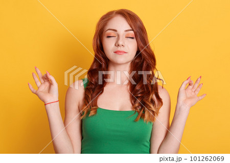 Good looking woman meditates, keeps both hands in okay gesture, keeps eyes closed, relaxing after work, practices yoga, wears green shirt, isolated on green wall. Good looking woman meditates, keeps both hands in okay gesture, keeps eyes closed, relaxing after work, practices yoga, wears green shirt, isolated on green wall. 101262069