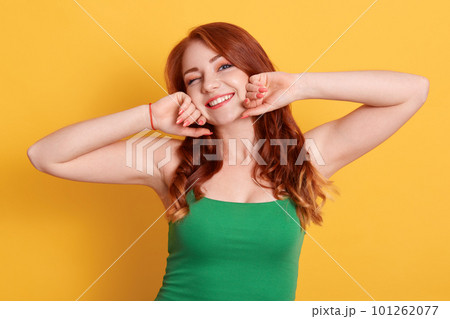 Beautiful smiling woman stretching and winking while looking directly at camera, wearing green sleeveless t shirt, red haired lady with pleasant appearance and positive emotions. 101262077