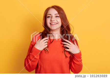 Cheerful gorgeous young woman wearing orange jumper, smiling happily while looking aside. Pretty girl excited joyful smile touching her red hair against yellow background. 101263089