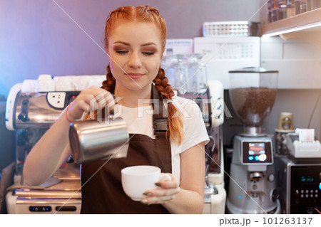 Red haired barista girl brews coffee from turk in coffee shop, concentrated woman working in cafe, lady in white t shirt and brown apron with coffee machine in background. Red haired barista girl brews coffee from turk in coffee shop, concentrated woman working in cafe, lady in white t shirt and brown apron with coffee machine in background. 101263137