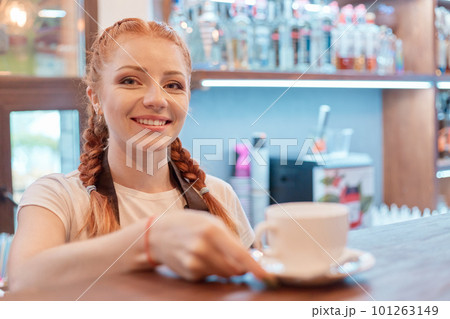 Young smiling woman standing at bar in coffee shop, barista posing at counter, offers cup of hot beverage for her client, red haired female working in cafe. Young smiling woman standing at bar in coffee shop, barista posing at counter, offers cup of hot beverage for her client, red haired female working in cafe. 101263149