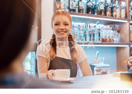 Happy young barista offering cup of coffee while smiling at camera or to her client, wearing white casual t shirt and brown apron, posing at counter. 101263150