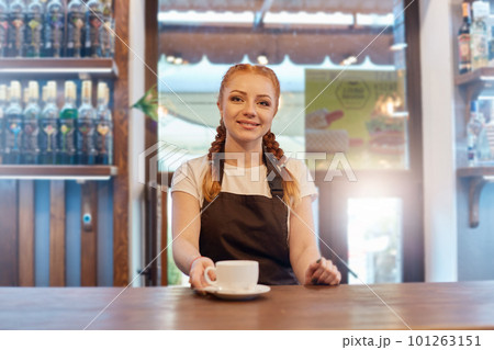Beautiful Caucasian rad haired barkeeper looks directly at camera offers hot coffee in white ceramic cup at coffee shop, barista posing near counter, dresses t shirt and apron. 101263151