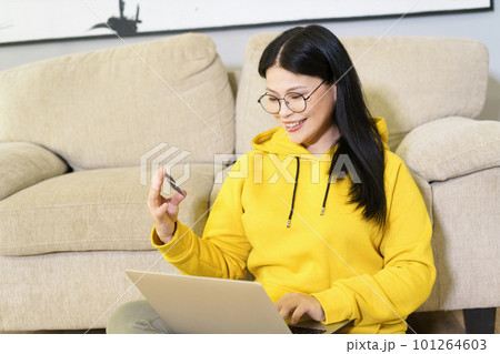 Asian woman comfortably shopping online from her home, with laptop on her lap and bank card in her hand. She is engaged in digital retail, using modern technology and the convenience of e-commerce to Asian woman comfortably shopping online from her home, with laptop on her lap and bank card in her hand. She is engaged in digital retail, using modern technology and the convenience of e-commerce to 101264603