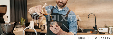 Smiling barista with stylish beard wearing apron poured filter coffee into a glass Smiling barista with stylish beard wearing apron poured filter coffee into a glass 101265135