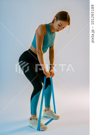 Strong woman in sportswear performs fitness exercises with resistance band on studio background Strong woman in sportswear performs fitness exercises with resistance band on studio background 101265189