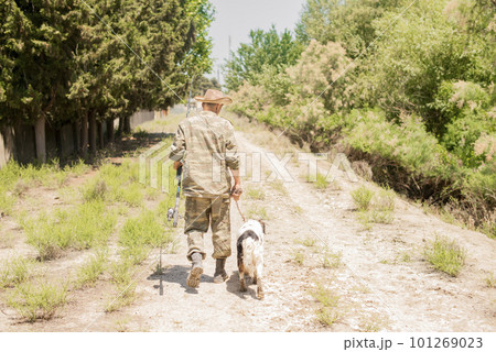 Unrecognizable, slender male fisherman in camo outfit walking down dirt road with obedient spotted dog in summer. Back view of adult guy with fishing pole going along bushy path. Concept of hobby. Unrecognizable, slender male fisherman in camo outfit walking down dirt road with obedient spotted dog in summer. Back view of adult guy with fishing pole going along bushy path. Concept of hobby. 101269023