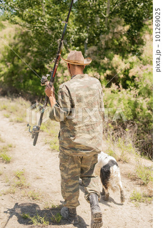 Unrecognizable fisherman in camouflage going on fishing with setter dog at daytime. Back view of slim fisher carrying big rod, while walking along path to lake outdoors. Concept of hobby, lifestyle. 101269025