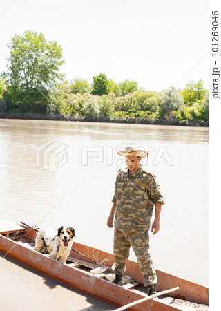 Concentrated male angler with faithful dog standing in old boat, going mooring after fishing in countryside. Front view of male fisherman enjoying trip on murky river. Concept of fishery, hobby. 101269046