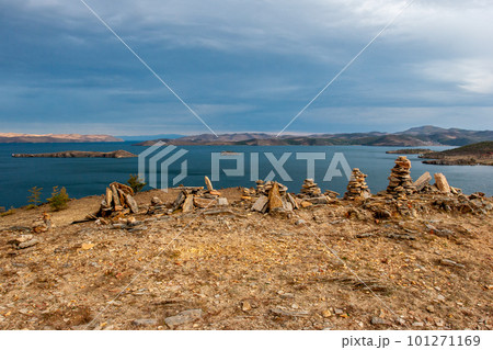 Stones piled by pyramids and a view of Lake Baikal. Hills beyond the lake. Several islands in the lake. Cloudy and gloomy. Horizontal. 101271169