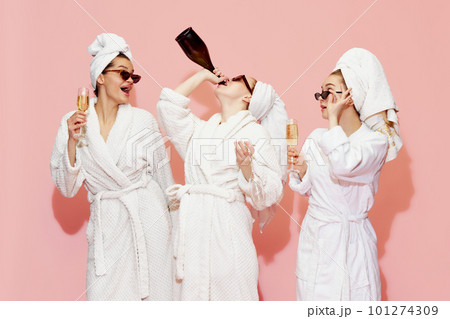 Spa meeting. Three beautiful girls in bathrobes drinking champagne, having fun against pink studio background. Celebration 101274309