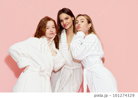 Portrait od three beautiful girls, friends in white bathrobes posing against pink studio background. Spa weekends, self-care 101274329