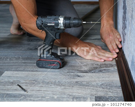 a man installs a floor wooden plinth made of dark lacquered wood in a room with gray textured linoleum using a screwdriver, holding the plank with his hands at the junction of wall and floor a man installs a floor wooden plinth made of dark lacquered wood in a room with gray textured linoleum using a screwdriver, holding the plank with his hands at the junction of wall and floor 101274740