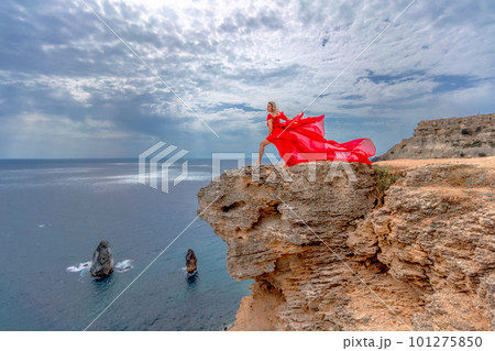 A woman in a red silk dress stands by the ocean, with mountains in the background, as her dress sways in the breeze. A woman in a red silk dress stands by the ocean, with mountains in the background, as her dress sways in the breeze. 101275850