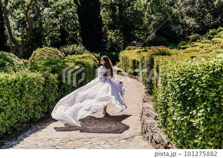 Brunette runs white dress park. A beautiful woman with long brown hair and a long white dress runs along the path along the beautiful bushes in the park, rear view 101275882