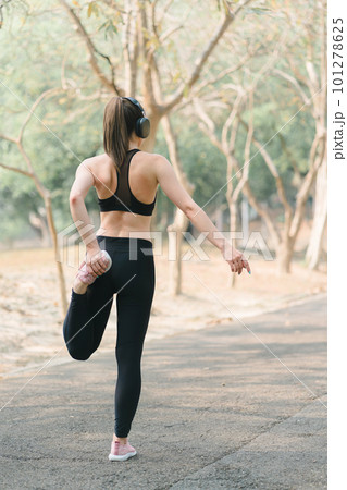 In an Autumn city park background, a stunning sportswoman in sportswear is smiling while stretching her body and performing flexibility exercises as a warm-up before a running workout. 101278625