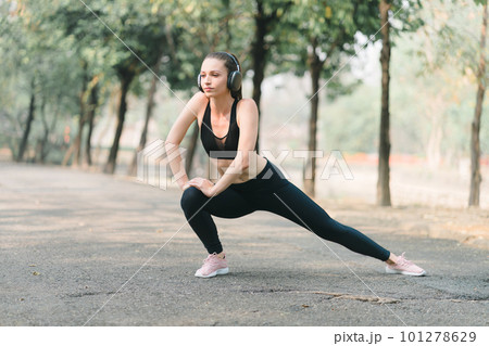 A beautiful sportswoman in sportswear is stretching her body with a smile on her face, while doing flexibility exercises to warm up before a running workout in Autumn city park background. A beautiful sportswoman in sportswear is stretching her body with a smile on her face, while doing flexibility exercises to warm up before a running workout in Autumn city park background. 101278629