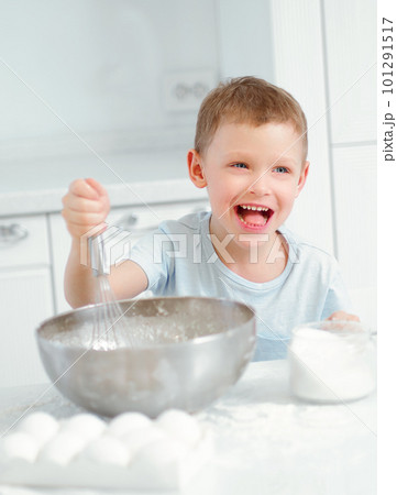 Child cooks food. Caucasian boy sits at table in kitchen and kneads dough in cup with hand whisk Child cooks food. Caucasian boy sits at table in kitchen and kneads dough in cup with hand whisk 101291517