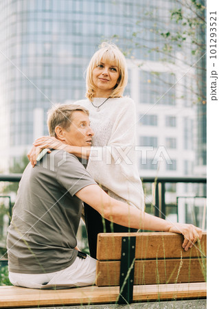 An adult mature happy romantic couple in love sitting on bench outdoors in city street park. A blonde caucasian man and woman spend time together. Senior wife and husband walking outside. 101293521