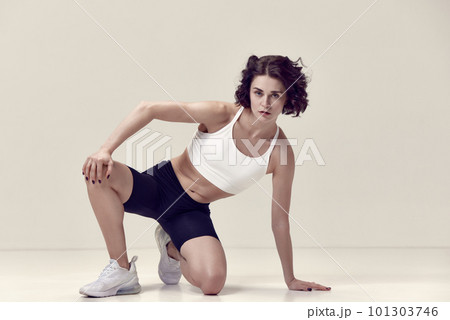 Pretty young woman with curly hair and fit, muscular, slim body in sportswear posing against light beige studio background Pretty young woman with curly hair and fit, muscular, slim body in sportswear posing against light beige studio background 101303746