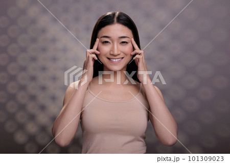 Skincare asian model showing eyes anti wrinkle massage procedure, touching temples and smiling. Natural beautiful young woman doing skin care treatment routine and looking at camera Skincare asian model showing eyes anti wrinkle massage procedure, touching temples and smiling. Natural beautiful young woman doing skin care treatment routine and looking at camera 101309023