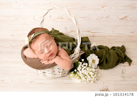 A newborn baby girl in a green floral headband sleeps in a wooden white basket with a green blanket, top view. On a light wooden background. 101314178