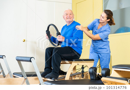 Healthcare professional helps an elderly man restore health with gymnastic ring in pilates class 101314776