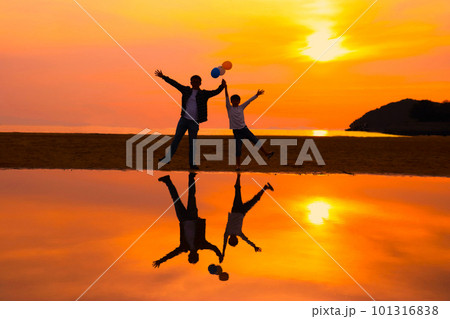 浜辺で夕日を背に風船を持って万歳をする父親と息子のシルエット 浜辺で夕日を背に風船を持って万歳をする父親と息子のシルエット 101316838